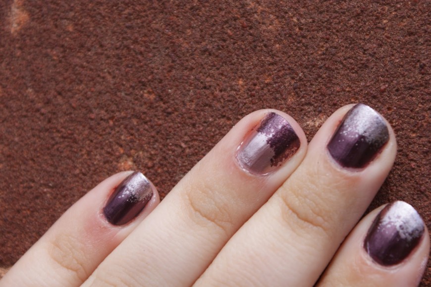 A hand with purple and silver ombre-painted nails rests against a textured rusty brown surface.