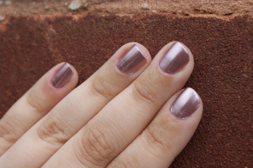 A hand with light skin is shown with neatly manicured nails painted in a metallic mauve polish. The fingers are lightly resting on a textured brick surface.