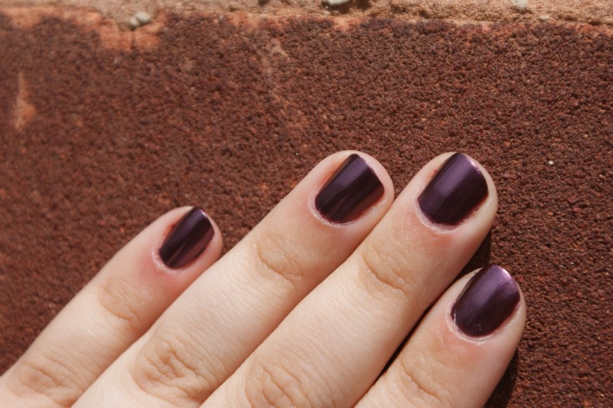 A hand with neatly manicured fingernails painted in dark purple polish rests against a textured brick surface under bright sunlight.