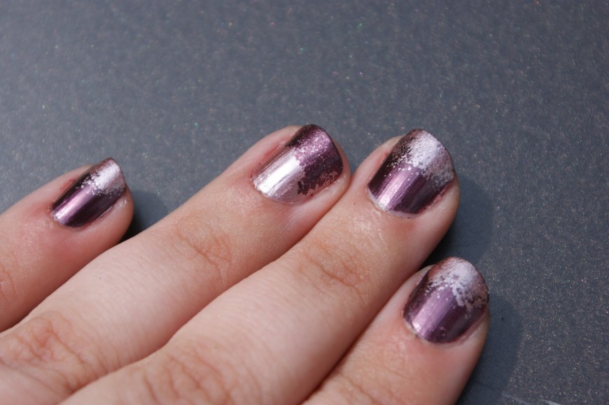 A hand with shiny, metallic purple ombre nails on a grey background. Each nail transitions from a deep purple at the base to a lighter, shimmering silver-pink at the tips.