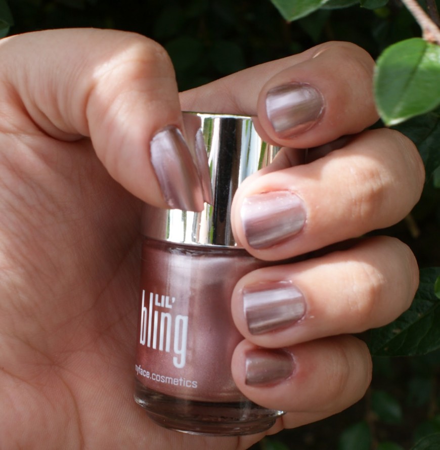 A hand with metallic mauve nail polish holds a bottle labeled bling by fyface.cosmetics. The nails are neatly manicured, and the background features green leaves.