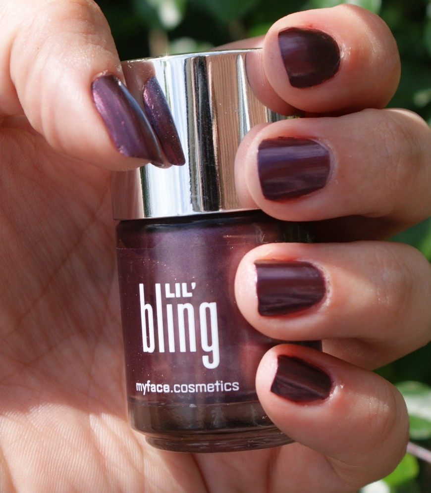 A hand with nails painted in a glossy, dark purple shade holds a bottle of Lil Bling nail polish by myface.cosmetics. The background features blurred greenery.