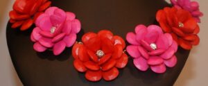 A necklace taking shape with alternating red and pink flower-shaped beads, each boasting a central gem. The elegant piece is displayed on a dark bust stand.