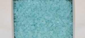 A close-up image showcases numerous small blue-green crystals, reminiscent of Mad Beauty products, filling a rectangular container with white edges. The crystals have an irregular shape, like coarse grains of salt or decorative stones.