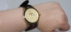 A wristwatch with a gold face and black and red striped fabric strap is on a persons wrist. The watch displays the day and date in a small window on the right side, showing WED and 9.