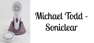 A Michael Todd Soniclear facial cleansing brush on a stand, with an additional brush head beside it. The background is white, and bold text on the right reads Michael Todd - Soniclear.