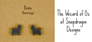 Two black earrings shaped like small dogs on brown cardboard. Text on the left reads Toto Earrings. Text on the right reads The Wizard of Oz at Snapdragon Designs.