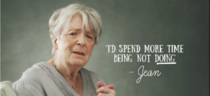 An elderly woman with short gray hair is seated, looking thoughtfully at the camera. The text next to her reads, Id spend more time being, not doing - Jean. The background is a plain, muted gray.