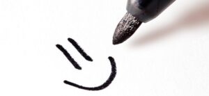 Close-up of a black marker drawing a simple smiley face on white paper. The face consists of two vertical lines representing eyes and a curved line for a smile.