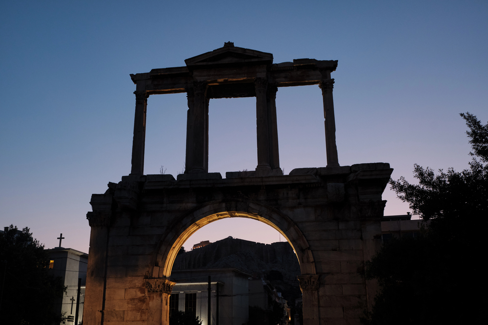 The stone ruins of Hadrian's Arch in Athens at dusk, with the illuminated Parthenon visible on the hill in the background.