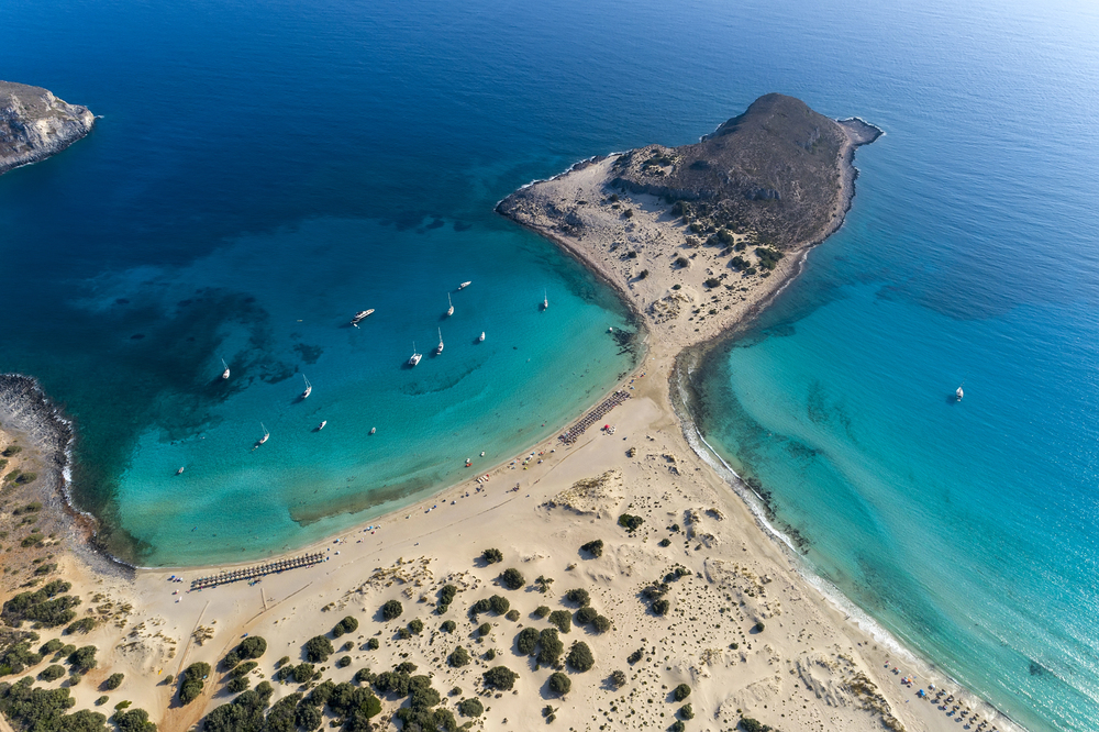 Aerial view of a sand spit connecting a rocky island to a mainland beach, surrounded by turquoise water with several small boats anchored in the bay.