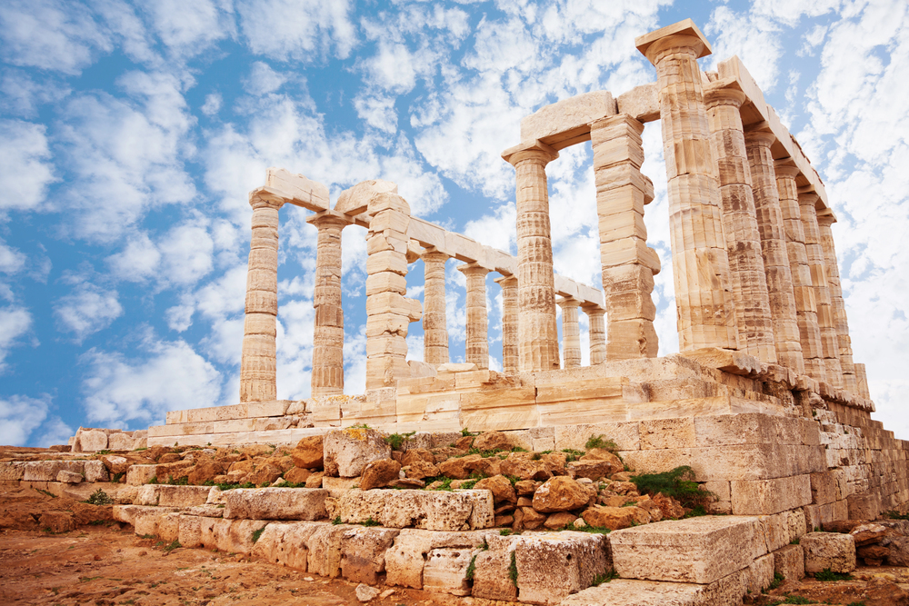 The ruins of an ancient Greek temple with stone columns against a bright blue sky with scattered clouds.