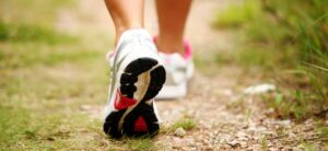 Close-up of a person walking on a gravel path, wearing sneakers with white, pink, and black accents. The focus is on the shoes, and the background is slightly blurred with green foliage.