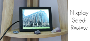 A digital photo frame displaying an image of a cathedral sits on a wooden shelf, surrounded by small stones. The text Nixplay Seed: Review is on the right.