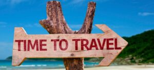 A wooden arrow sign with the words Time to Travel points right. It is mounted on a piece of wood, set against a backdrop of a sandy beach with a blue sky and green hills in the distance.