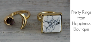 Two gold rings are displayed on a wooden surface. One features a crescent shape with a small purple stone, and the other has a large square setting with a white marbled pattern. Text on the right reads Pretty Rings from Happiness Boutique.