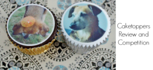 Two cupcakes with edible dog images, one in a gold wrapper and the other in a silver wrapper, are placed on a patterned surface. Text reads: Caketoppers Review and Competition.