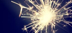 A bright, sparkling firework against a dark blue background. The light bursts radiate outward, creating a starburst effect with glowing lines and scattered sparkles.