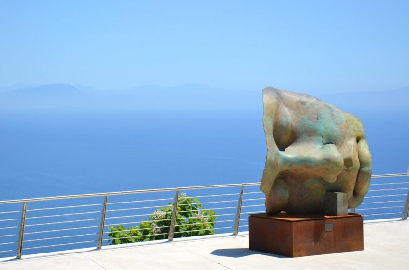 Bronze sculpture resembling a stylized head with abstract features, set on a stone pedestal. It overlooks a vast expanse of blue ocean under a clear sky, with a metal railing and greenery visible in the background.