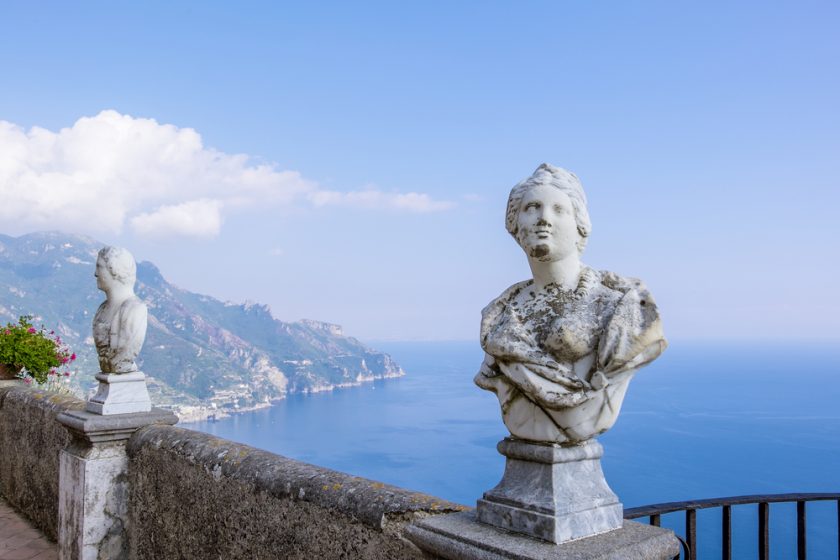 Marble busts on a terrace overlooking the Amalfi Coast, with clear blue sky and sea. The scenic view includes distant cliffs and lush greenery.