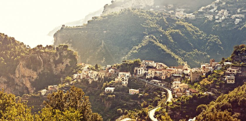 A picturesque hillside village with rustic buildings nestled among lush green mountains under a hazy sky. A winding road curves through the landscape, leading to the quaint settlement. Mist softly settles in the distance, blending with the terrain.