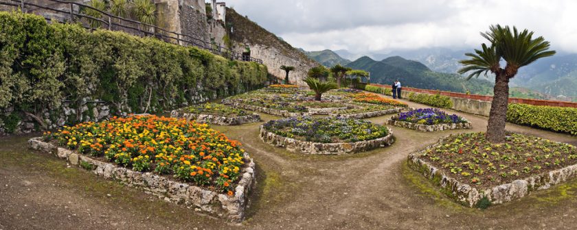 A panoramic view of a landscaped garden with neatly arranged flower beds in various shapes, featuring orange, yellow, and purple blooms. A dirt path winds through the garden. Mountains and cloudy skies are visible in the background.