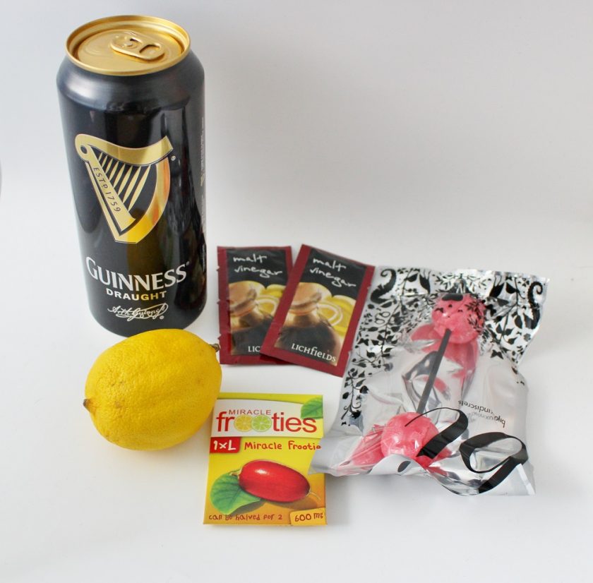 JustUsBox Date Night Box miracle berry frooties alongside 2 packets of vinegar, double ended lollipop, lemon and can of Guinness