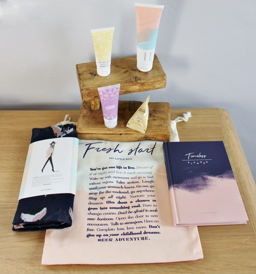 My Little Box January 2017 over view photo of the whole box; bag in the centre with the snood to the left and planner to the right. Beauty items behind the bag are on tiny wooden shelves.