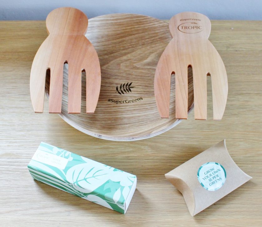 Tropic Skin Care SUPER GREENS along side a bottle of the SUPER GREENS Tropic Skin Care sent me a wooden salad bowl and servers, the bottom of the bowl includes the words SUPER GREENS alongside a picture of some leaves, to the bottom right of the bowl is a small packet of seed bombs, to grow my own greens and to the right is the full packaged in the cardboard SUPER GREENS bottle, the cardboard outer uses different greens to make a leaf pattern.