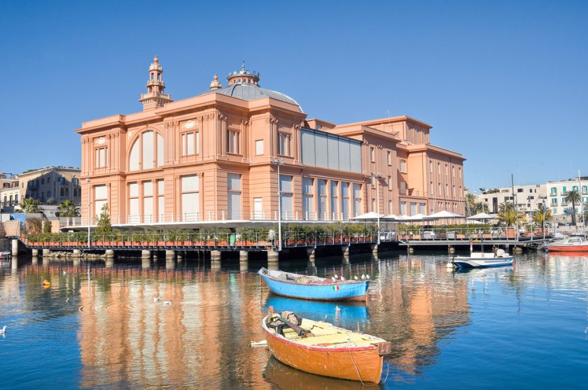 Waterfront scene with a large, pinkish building featuring domes. In the foreground, three small, colorful boats float on calm water, reflecting the building and boats. Clear blue sky in the background.