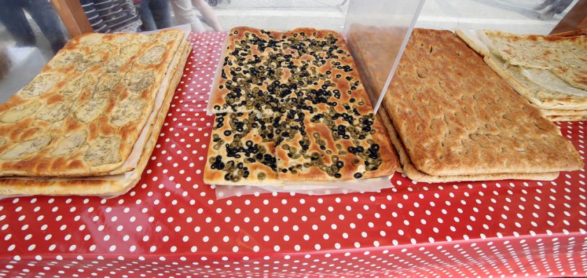 Three large rectangular focaccias displayed on a red polka dot tablecloth. The left one has herbs, the middle one is topped with black olives, and the right one is plain. The focaccias are behind a clear divider.