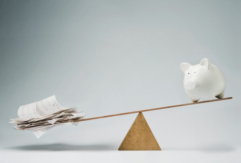 A seesaw balances a stack of receipts on one end and a white piggy bank on the other. The receipts outweigh the piggy bank, causing the seesaw to tilt. The background is a plain, light gray.