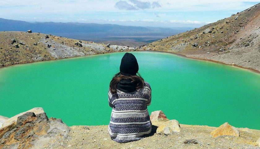 Person in a patterned sweater and black hat sits on a rocky edge, gazing at a vibrant turquoise lake surrounded by barren hills under a cloudy sky.