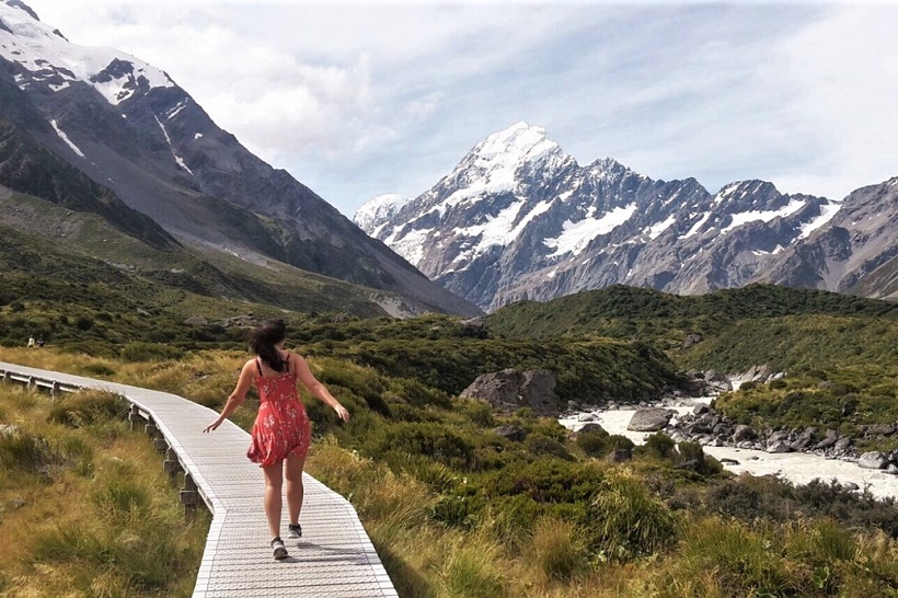 A person in a red dress walks on a wooden boardwalk surrounded by green grass and shrubs, with snow-capped mountains and a cloudy sky in the background.
