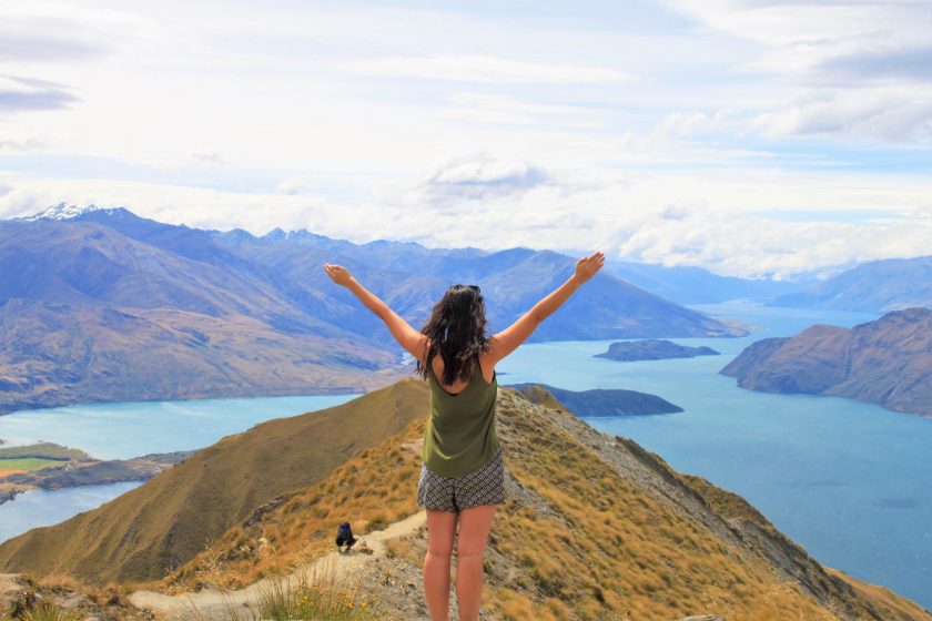 A person with long hair stands on a grassy mountain peak, arms raised in celebration, overlooking a vast landscape of mountains and a blue lake under a partly cloudy sky.