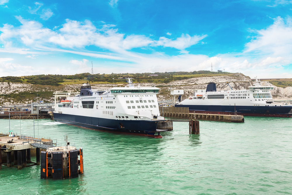 Two large white and blue ferries docked at a port with green water, under a bright blue sky. White cliffs and greenery are visible in the background.