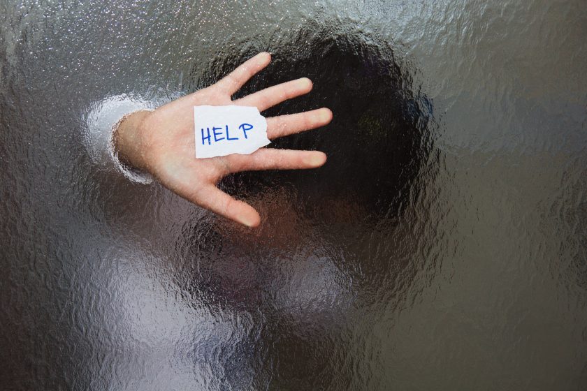 A person presses their hand against frosted glass, holding a small piece of paper with HELP written on it. Their face and body are blurred behind the textured surface.