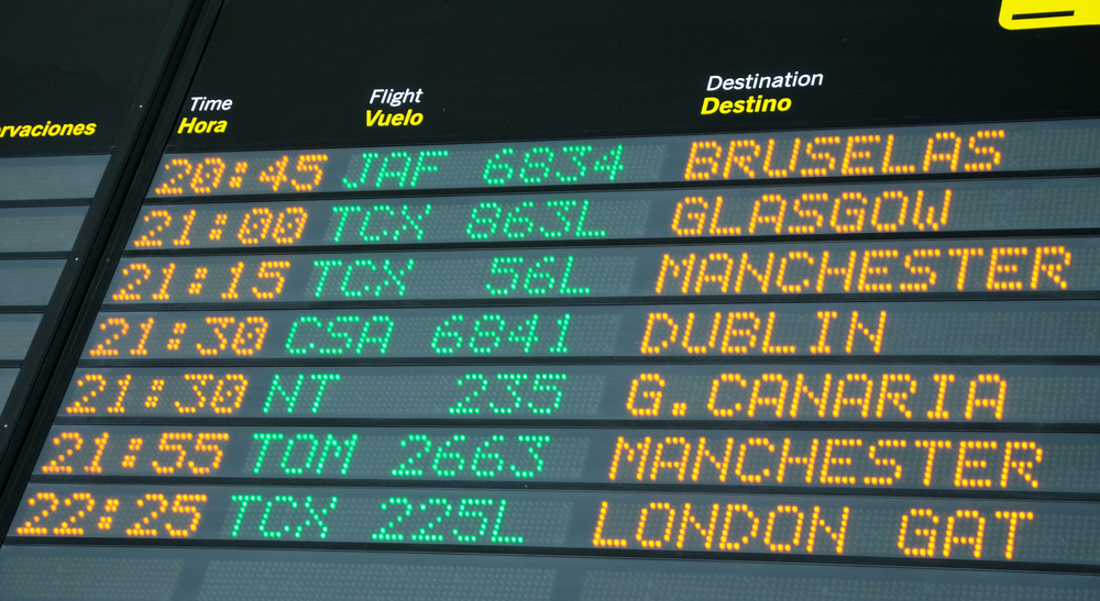 An airport departure board displays flight times, numbers, and destinations including Brussels, Glasgow, Manchester, Dublin, Gran Canaria, and London Gatwick, with text in Spanish and English.