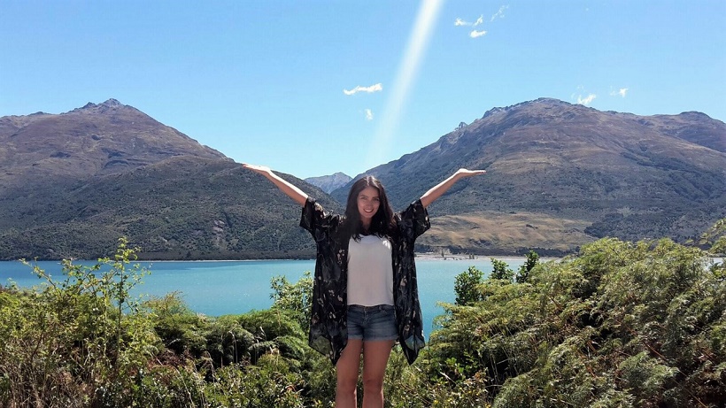A woman stands smiling with arms raised in front of a scenic backdrop of mountains and a lake. She is wearing a black floral kimono, white top, and denim shorts. Green shrubs are in the foreground, and the sky is clear and blue.