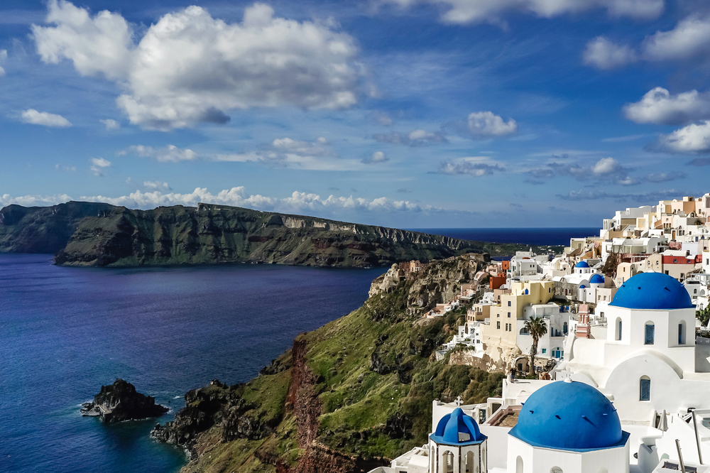 White buildings with blue domes overlook the deep blue sea and rugged coastline in Santorini In The Winter, Greece, under a sky dotted with fluffy clouds. A green island is visible across the water.