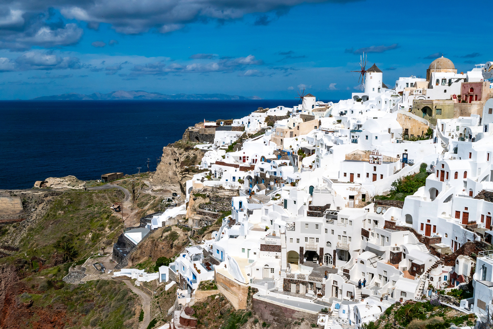 Whitewashed buildings with blue rooftops cluster on a cliffside overlooking the deep blue sea in Santorini, Greece, under a clear sky, with a traditional windmill visible—Santorini in the winter feels especially serene and magical.
