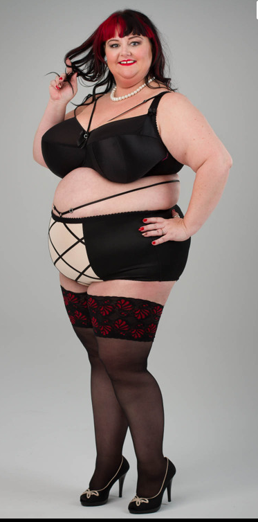A woman poses confidently in black lingerie with beige accents and black stockings featuring red patterns. She has shoulder-length dark hair with red highlights, wears pearl jewelry, and black high heels, standing against a gray background.