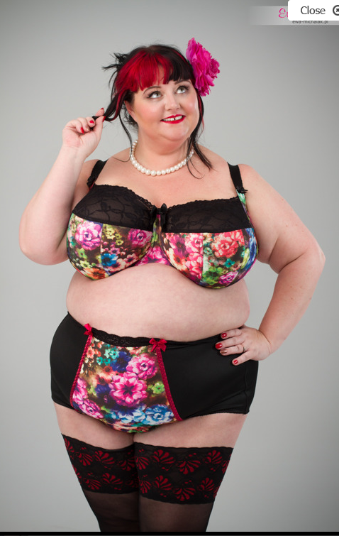 A woman poses confidently in colorful floral-patterned lingerie, wearing a pearl necklace and a pink flower in her hair. She smiles with her hand playfully touching her hair, standing against a plain gray background.