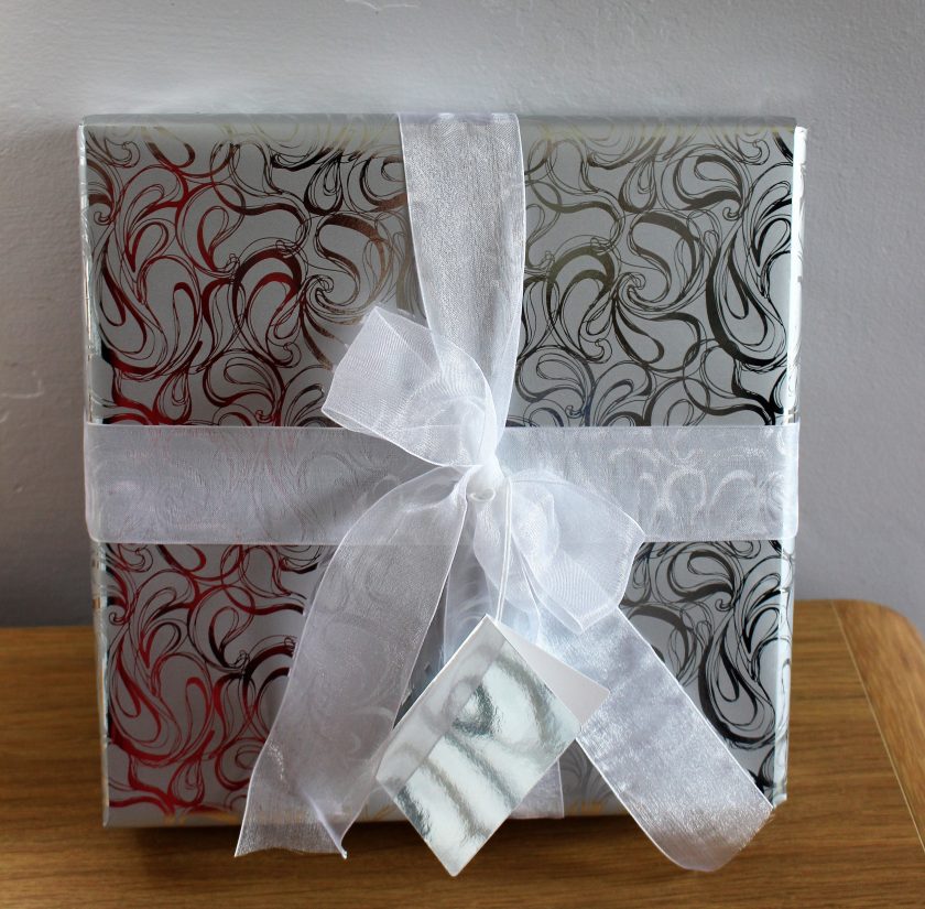 A gift wrapped in shiny, swirled-patterned paper with hues of red, black, and gold. Tied with a large, sheer white ribbon, it includes a matching gift tag. The present rests on a wooden surface against a plain background.