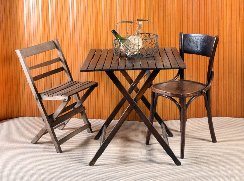 Furniture Still Life of Folding Table with Mismatched Chairs and Wire Basket with Bottles in Room with Beige Carpet and Wood Wall Paneling