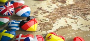 Heart-shaped pins with different national flags, including Germany, Greece, Sweden, and Spain, are placed on a vintage map of Europe. The map highlights regions like the Bay of Biscay and cities such as Paris and Madrid.