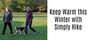 A man and woman walk a dog on a grassy area covered with fallen leaves. The text on the right says, Keep Warm this Winter with Simply Hike. Trees are visible in the background.