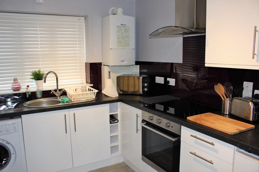At Owl Apartments, discover a modern kitchen featuring white cabinets and black countertops. Equipped with an electric stove, oven, microwave, toaster, and washing machine. A cutting board and utensils adorn the counter alongside a small plant and dish rack near the sink with window blinds.
