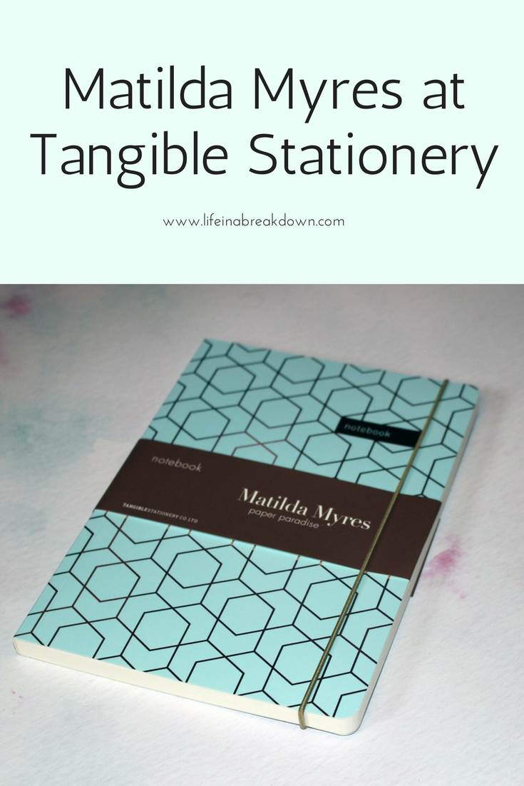 A notebook with a geometric pattern and a brown label that reads Matilda Myres paper paradise rests on a white surface. An elastic band keeps it secure. The text above proudly announces, Discover Matilda Myres at Tangible Stationery.