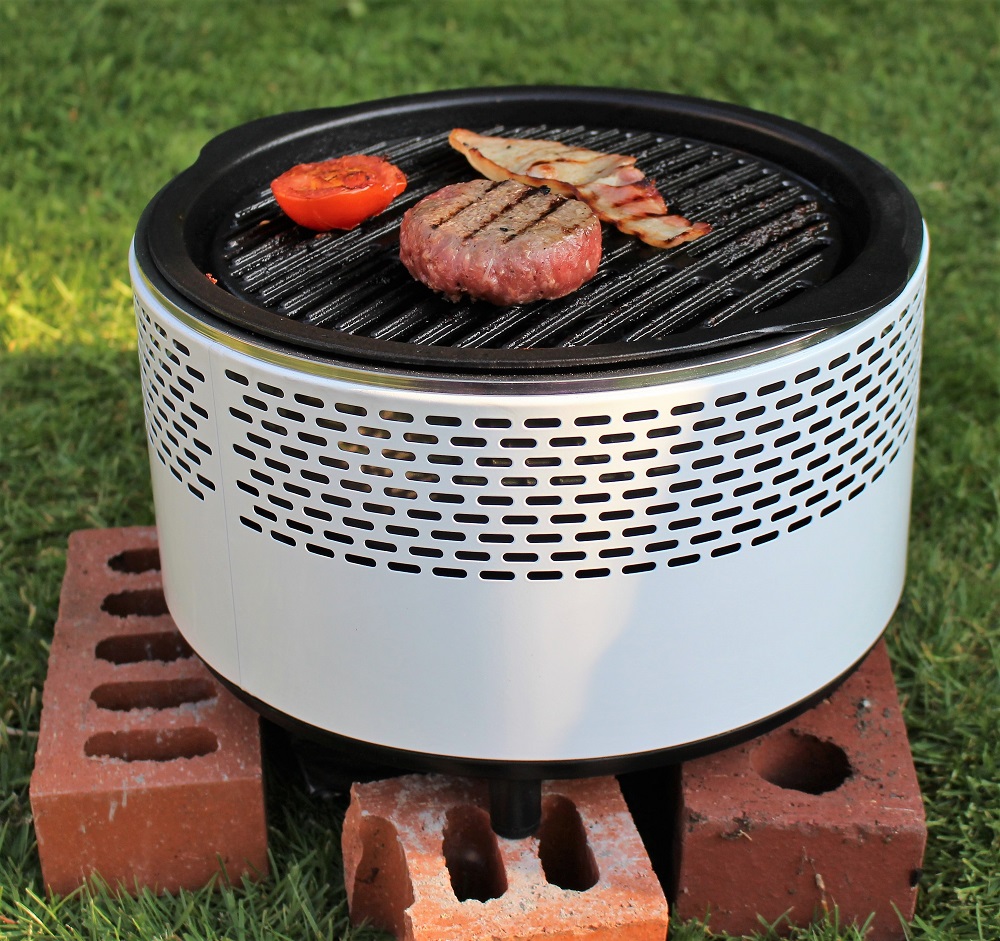 A smokeless BBQ grill sits on a lawn, perched on bricks, sizzling with a burger patty, sliced tomato, and crispy bacon. The round metal design features ventilation holes that make it ideal for smoke-free outdoor cooking.